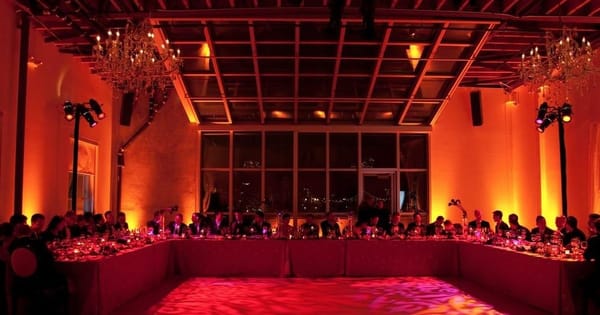Long U-shaped dinner table under a glass skylight roof, with crystal chandeliers, amber uplighting and a patterned light projection on the floor