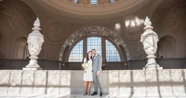 Bride and groom standing beneath the dome inside San Francisco City Hall, between two carved stone urns