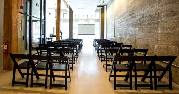 Rows of black folding chairs facing a projection screen in a long event room with a concrete wall, timber posts and a glass storefront