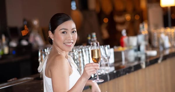 Smiling woman with a sleek low bun and soft glam makeup in a white one-shoulder dress, raising a glass of sparkling wine at a bar