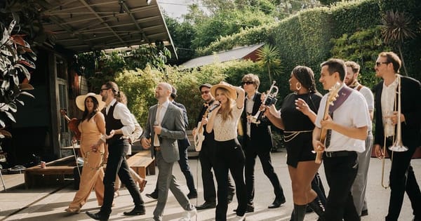 Band members in suits and hats walk across a sunny garden patio carrying a trombone and guitars, with tall hedges behind