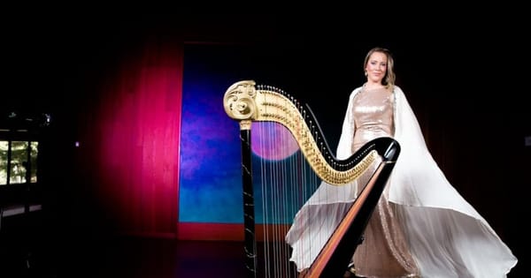 Harpist in a sequined gown and flowing white cape standing beside a gold and black concert harp on a dark stage
