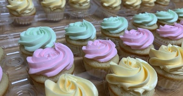 Rows of vanilla cupcakes with swirled pastel pink, mint green and pale yellow frosting in clear plastic trays