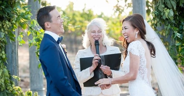 Laughing bride and groom holding hands in front of a smiling officiant with a microphone and black folder, framed by leafy vines outdoors