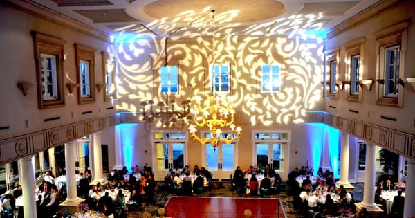 Reception ballroom with white scroll patterns projected across the walls and ceiling, blue uplights between the columns and guests at round tables