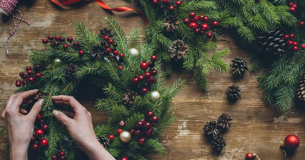 Hands arranging a fresh fir wreath with red berries, pine cones and small ornaments on a wooden table, with red ribbon and twine nearby